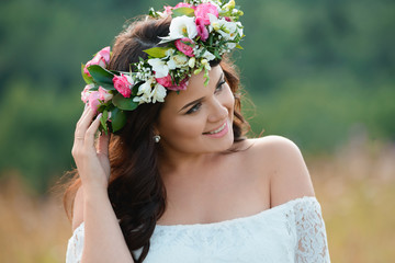 Obraz premium Smiling brunette girl with a wreath of flowers. Young woman with long hair in a white lace dress. Close-up female portrait