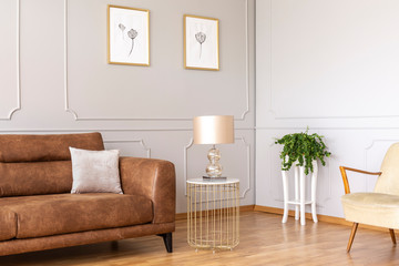 Gold lamp on table next to brown leather settee in grey loft interior with posters and plant. Real...