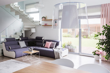 Wooden table next to corner sofa with pillows in white apartment interior with window. Real photo