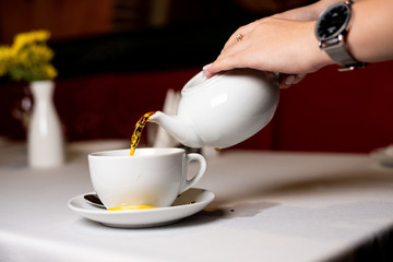 female hands pour white teapot tea into a cup that stands on the table