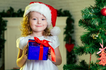Close-up portrait of a blonde curls little girl in santa claus hat with a gift dreaming about christmas miracles standing next to the christmas tree and fireplace bright interior of the house