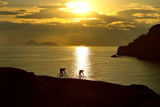 Two mountain bikers cycling on a rocky coast, back light at sunset, Red Beach, Matala, Crete, Greece, Europe