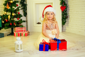 Obraz na płótnie Canvas Cute little girl in dress Santa Claus hat curly blondу hair wide open eyes holding a lot of gifts box at home near a Christmas tree with gifts and garlands and a decorated fireplace .new Year morning