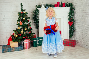 Cute little girl in a nice dress with long curly blondу hair holding a box with a gift at home near a Christmas tree with gifts and garlands and a decorated fireplace .new Year morning