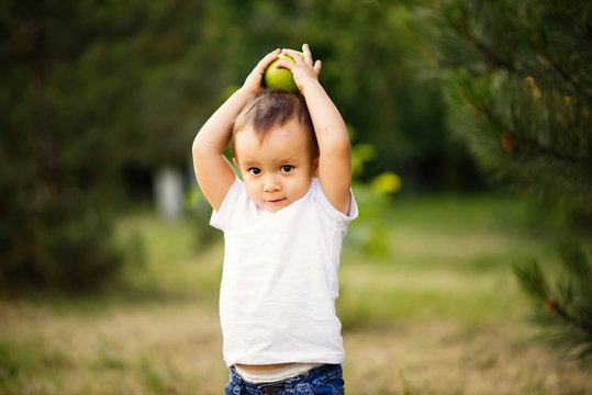 .Little Child In White Shirt Holding A Green Apple On His Head. Toddler Boy With An Apple Outdoor Near Pine Tree.