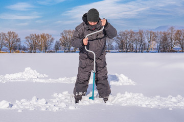 Fototapeta na wymiar Winter fishing concept. Fisherman in action, making the hole in snowy ice at lake by ice screw
