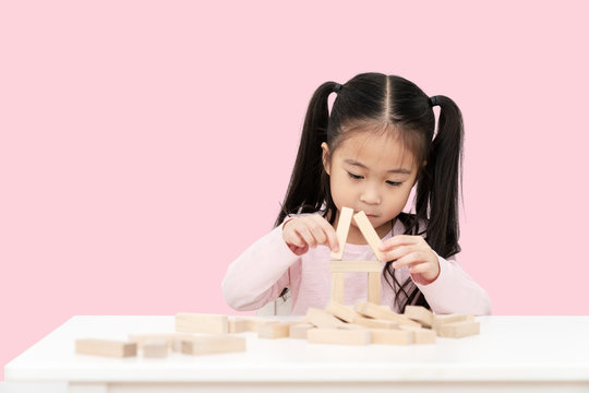 Young little cute asian girl build a house from wooden block construction, wood toy, jenga house on desk in isolated pink background. Asia children play and learn creative concept with copy space.