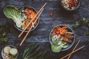 Delicious asian bowls with rice noodles, vegetables and tofu on wooden background. Top view