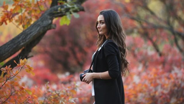 A charming young girl photographer shoots the autumn landscape with a digital SLR camera. A brunette with a beautiful smile in the autumn garden takes pictures of trees with red leaves.