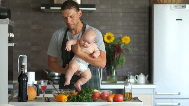 Young father in an apron holds a six month baby in his arms. The interior of the modern kitchen, on the table are fresh bright vegetables. Father's day