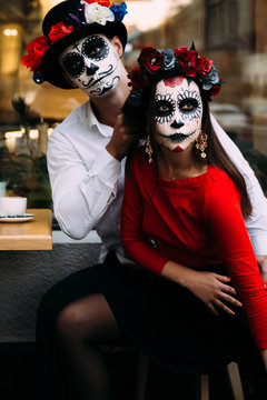 A couple, wearing skull make-up for. All souls day. Boy and girl sugar skull makeup.painted for halloween sit in a cafe. dead in the city. zombie walk.