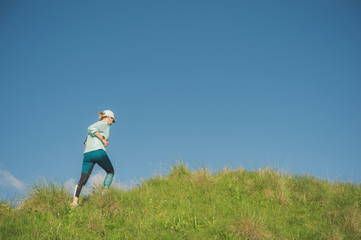 Attractive sports girl in a cap and headphones jogging through the grass in a picturesque place up the hill against the blue sky. Workout outdoors