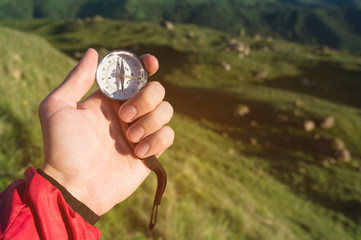 Man searching direction with a compass in hand in the summer mountains. View point on the background of green hills and grass. Direction Search