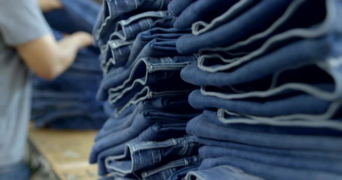 Focus on piles of folded jeans with unidentifiable male garment industry worker sorting clothes in background at warehouse in Los Angeles. Slow motion hand-held 4K recorded 60fps