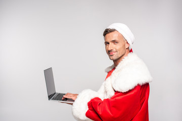 handsome man in santa costume using laptop and smiling at camera isolated on grey