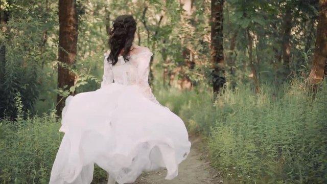 cute slender girl, dressed in a long white vintage transparent dress with floral patterns and a long waving train, runs along a narrow path in a forest. a boho style for runaway bride