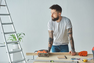 handsome bearded man leaning at wooden table and looking away during house repair