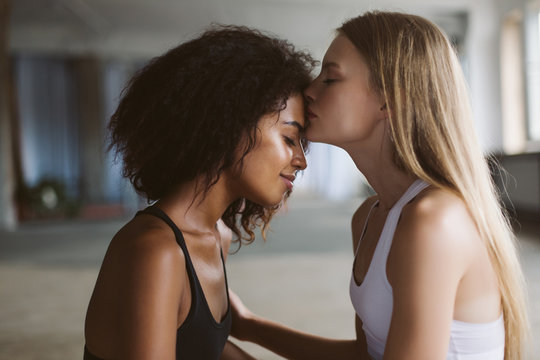 Close up young woman with blond hair thoughtfully kissing in forehead smiling african american woman with dark curly hair while spending time together at home