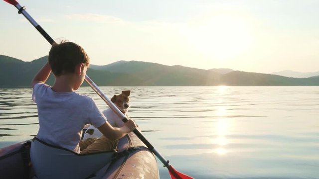 Slow motion of a happy boy with his dog Jack Russell Terrier paddling an inflatable kayak on the water of a large mountain lake against the backdrop of beautiful orange sunset. Family sports vacation