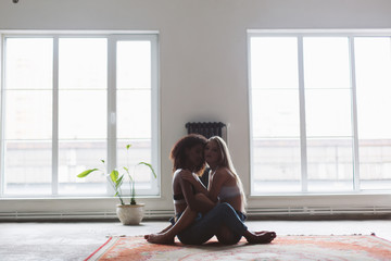 Young attractive woman with blond hair and african american woman with dark curly hair sitting on carpet while dreamily looking in camera spending time together at home with big windows on background