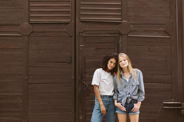 Young beautiful african american woman with dark curly hair in T-shirt and jeans and woman with blond hair in shirt and denim shorts dreamily looking in camera with brown wall on background