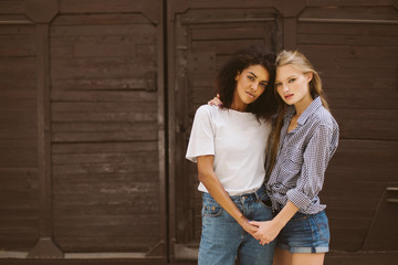 Young pretty african american woman with dark curly hair in T-shirt and jeans and attractive woman with blond hair in shirt and denim shorts dreamily looking in camera while spending time together