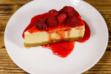 Piece of tasty New York cheesecake with raspberries and raspberry jam in a white plate on wooden table