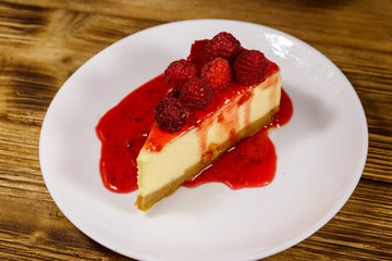 Piece of tasty New York cheesecake with raspberries and raspberry jam in a white plate on wooden table