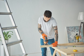 handsome bearded tattooed man using electric jigsaw during house repair
