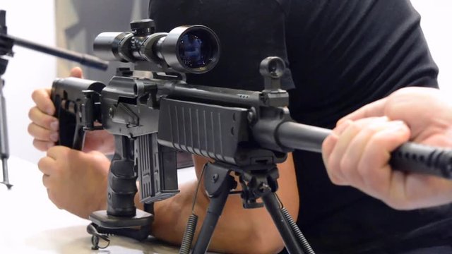 Several large-caliber weapons on the table. Person looks at the handle of a rifle standing on the surface.