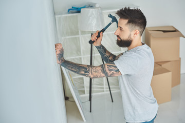 side view of bearded young man hammering nail at wall in new house