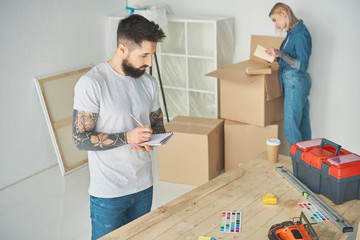 bearded man taking notes and young woman unpacking cardboard boxes in new house