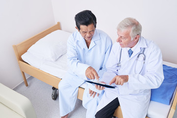 From above shot of elderly medical practitioner and Asian patient pointing at modern tablet while sitting on bed in ward