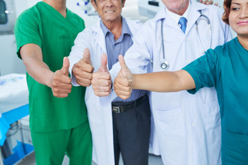 Unrecognizable medical practitioners and surgeons showing thumb-up gesture to camera while standing in operating theatre