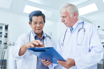 Two senior medical practitioner having discussion about information on clipboard while standing in operating theatre together