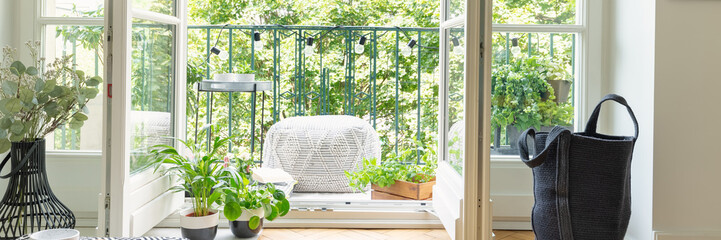 Panoramic view of open balcony door, black bag, green plants and light grey pouf - obrazy, fototapety, plakaty