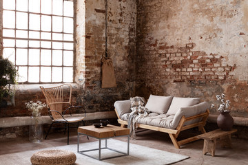Armchair and beige sofa in industrial living room interior with wooden table, pouf and window. Real...