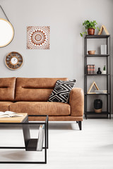 Patterned pillow on leather couch in grey ethno apartment interior with table and poster. Real photo