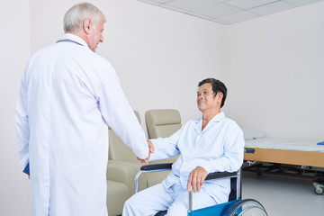 Elderly man in doctor uniform shaking hand of Asian male patient on wheelchair in modern ward of...
