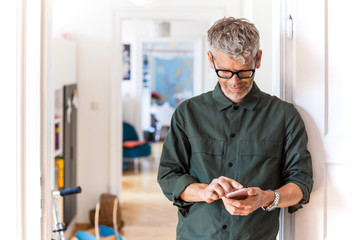 Mature man leaning against door case at home using cell phone