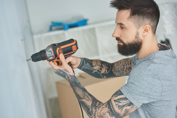 tattooed man doing hole in wall with screw gun in new apartment, inexperienced millennial concept