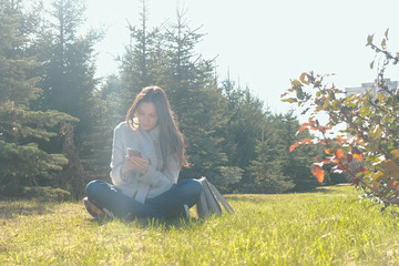 Young beautiful brunette woman sits on grass in spring park and types a message in mobile phone.