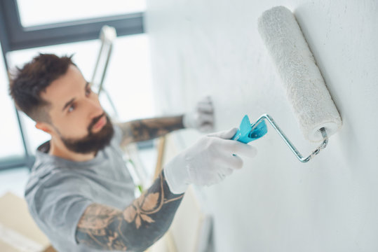 selective focus of tattooed repairman with roll paint brush painting wall in new apartment