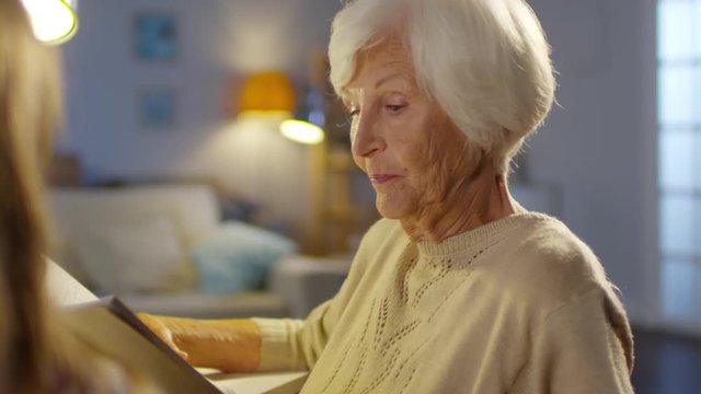Beautiful grandmother sitting at table in the living room and reading a book aloud to child