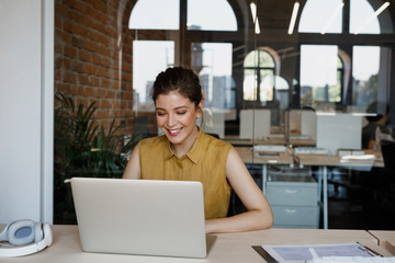 Pretty Caucasian smiling businesswoman sitting at her modern office and typing on her laptop.