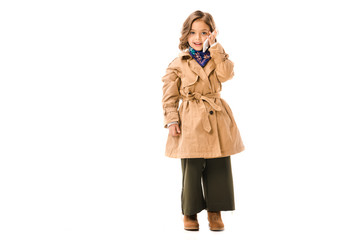 beautiful little child in trench coat talking by phone and looking at cameraisolated on white