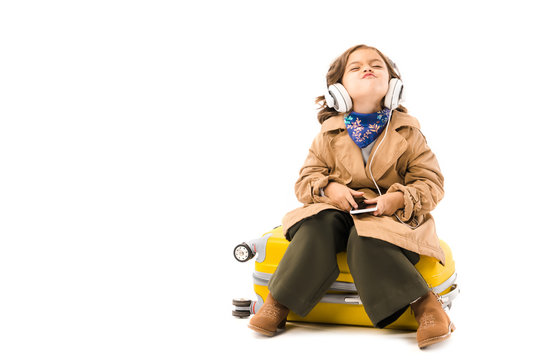 grimacing little child in trench coat listening music with headphones and sitting on yellow suitcase isolated on white