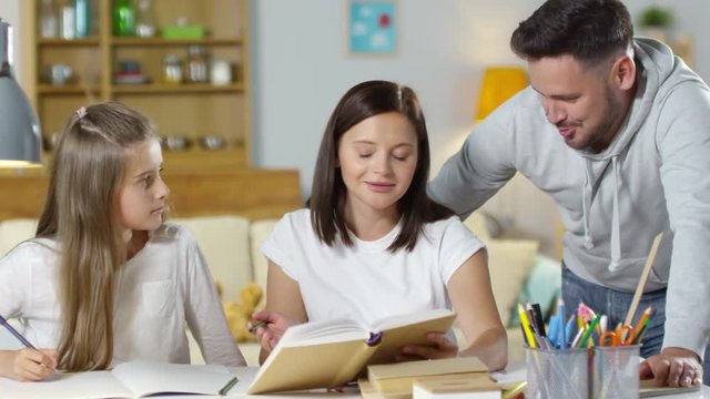 Beautiful mother smiling and talking with husband and young daughter while doing homework together at table in the living room