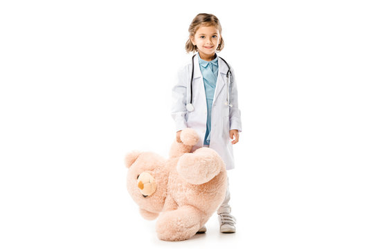 kid dressed in doctors white coat with stethoscope holding big teddy bear isolated on white