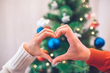 Nice picture of people's hands making hearts in front of Christmas tree with toys. There is some lights as well. Poeple wears red and white sweaters.